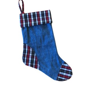 May include: A denim Christmas stocking with a red, black, and white plaid cuff, heel, and accent. The stocking has a loop for hanging and is made of blue denim fabric.