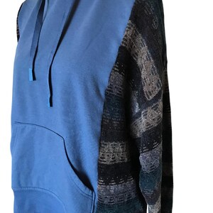 May include: A blue hooded sweatshirt featuring a solid blue front panel with a matching drawstring hood and a large kangaroo pocket. The sleeves and side panels are constructed from a textured knit fabric with a dark multi-colored geometric pattern.