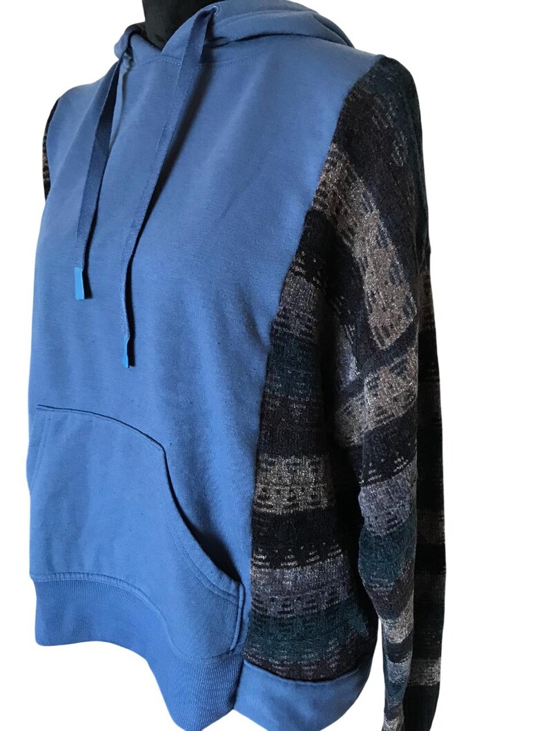 May include: A blue hooded sweatshirt featuring a solid blue front panel with a matching drawstring hood and a large kangaroo pocket. The sleeves and side panels are constructed from a textured knit fabric with a dark multi-colored geometric pattern.