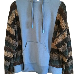 May include: A long-sleeve hoodie featuring a solid light blue center panel with a matching hood and drawstring, contrasted by sleeves and side panels patterned with horizontal stripes in shades of brown, dark green, and tan with a ribbed hem.