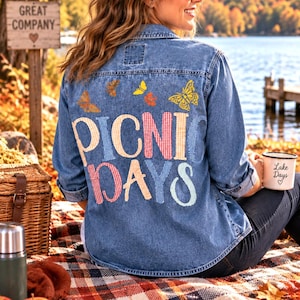 May include: A blue denim button-down shirt featuring the text PICNIC DAYS in colorful gingham patterns on the back, accented with small butterfly graphics. The shirt has long sleeves, a collar, and a classic denim texture with visible stitching.