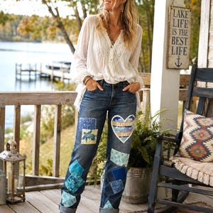 May include: A white long-sleeved blouse with lace detailing and dark blue denim jeans featuring various fabric patches in shades of blue and a sequined heart-shaped patch on the thigh with the text Slay The Day printed in white letters.