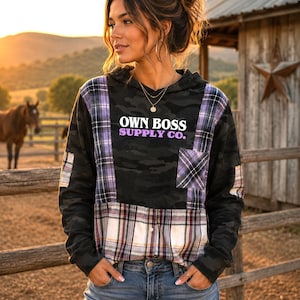 May include: A long-sleeve hooded sweatshirt featuring a black camouflage pattern combined with purple and white plaid fabric panels. The chest displays the white and purple text OWN BOSS SUPPLY CO. It includes a plaid chest pocket and plaid sleeve accents.
