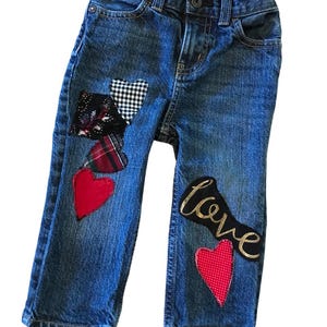 May include: Blue denim jeans with patches of red, black, and white fabric. The patches are in the shape of hearts and one patch has the word "love" written in gold glitter.