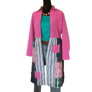 May include: A long, patchwork coat with a pink top and a patchwork bottom. The top is a solid pink color with a button-down design. The bottom is made of various fabric patterns, including stripes and plaid. A teal shirt and jeans are visible underneath.