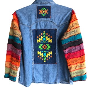 May include: A blue denim jacket with colorful patterned fabric sewn onto the sleeves and back. The back features two large geometric designs in yellow, orange, green, and blue.