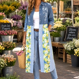 May include: A long-sleeved denim shirt dress featuring a blue and white gingham pattern with yellow and white floral panels, worn over light blue jeans with a brown belt and brown sandals, paired with a woven straw basket bag.