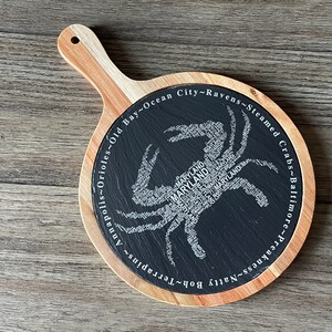 May include: A round wooden serving board with a black slate center. The slate features a crab design made of the word "Maryland" and other Maryland-related words. The board has a natural wood handle.