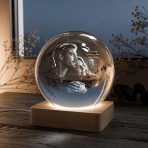 Op de afbeelding: Een heldere glazen bol met een zwart-wit afbeelding van een stel, rustend op een houten voet. De bol heeft de tekst "journey together, a love story etched in time" op het oppervlak. De basis wordt verlicht met een warme gloed.