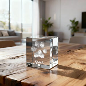 May include: A clear glass cube featuring a paw print design etched within. The word "Spark" is inscribed beneath the paw print, accompanied by a small heart symbol. The cube rests on a wooden table.