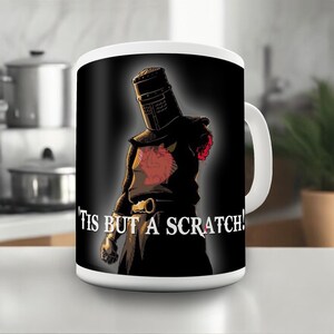 May include: White ceramic mug with a black background featuring a cartoon illustration of a knight in black armour with a red cross on his chest. The knight is missing his left arm and is saying "Tis but a scratch!"