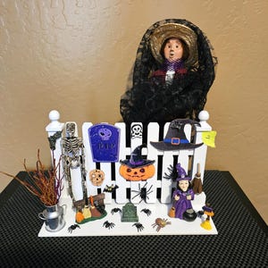 May include: A Halloween-themed decorative display featuring a white picket fence with various miniature figurines. Includes a skeleton, witch, pumpkin, and a doll wearing a black lace veil. The display is adorned with spiders and other spooky elements.