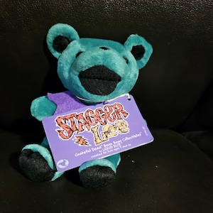 May include: A teal plush bear with black accents, wearing a purple vest. The bear has a tag that reads "Stagger Lee" with the words "Grateful Dead" and "Liquid Blue" printed on it. The bear is a collectible item.