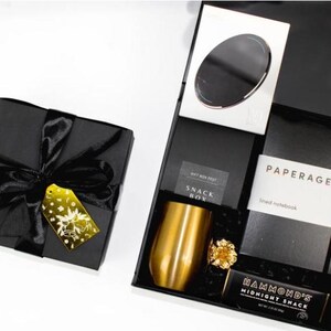 May include: A black gift box with a black satin ribbon and a gold gift tag. The box is filled with a variety of items, including a gold tumbler, a black notebook with the text "PAPERAGE lined notebook", a black box with the text "SNACK BOX", and a chocolate bar with the text "HAMMOND'S MIDNIGHT SNACK".