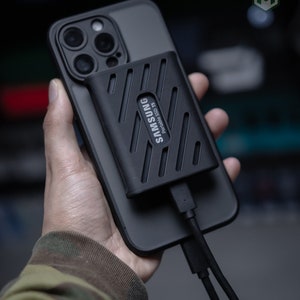 Puede incluir: Un disco duro externo negro Samsung Portable SSD T5 conectado a un iPhone negro con un cable USB-C.