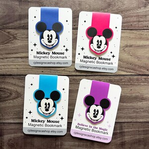 May include: Four magnetic bookmarks featuring Mickey Mouse heads on white backing cards. The bookmarks are blue, pink, light blue, and purple. Each card displays the text Mickey Mouse Magnetic Bookmark or Believe in the Magic Magnetic Bookmark.