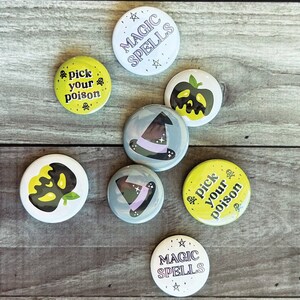 Könnte beinhalten: Sieben runde Knöpfe mit verschiedenen Halloween-Motiven. Die Knöpfe zeigen Bilder eines Hexenhutes, eines Kürbisses und den Text "Pick your poison" und "Magic Spells".