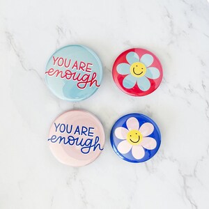 May include: Four round pins with a positive message. Two pins are light blue with the text "You are enough" in red and white. Two pins are blue with a white flower with a yellow smiley face.