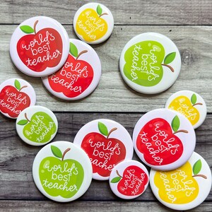 May include: Assortment of round, colorful buttons with the text "world's best teacher" in white lettering. The buttons feature red, green, and yellow apples with leaves.