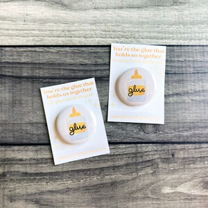 May include: Two white round magnets with a glue bottle design and the word "glue" in a handwritten font. The magnets are on white cards with the text "You're the glue that holds us together" and the shop's URL. The background is a gray wooden surface.