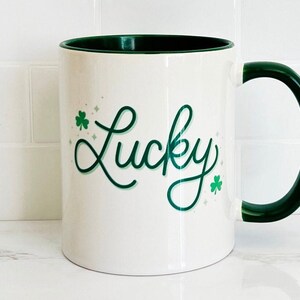 May include: White ceramic mug with a green rim and handle. The mug has a green shamrock design and the word "Lucky" written in green cursive.