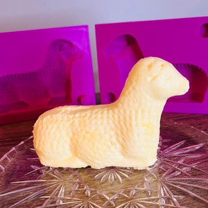 May include: A yellow butter lamb mold with a detailed textured surface. The mold is sitting on a clear glass plate with a decorative pattern. Two purple silicone molds are in the background.