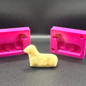 Pode incluir: Um doce amarelo em forma de cordeiro ao lado de dois moldes rosa brilhante. Os moldes são retangulares e apresentam um design de cão dachshund. O fundo é uma superfície cinza escuro.