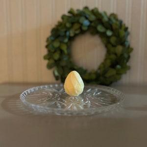 May include: A single, pale yellow, egg-shaped object sits on a clear glass plate with a decorative pattern. The object is in the centre of the plate, and the plate is on a light grey surface. A green wreath is in the background.