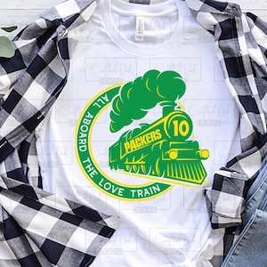 May include: A white t-shirt with a green and yellow train design. The train has the word "Packers" and the number "10" on it. The design is surrounded by a circle with the text "All Aboard The Love Train".