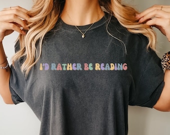 Puede incluir: Camiseta gris oscuro con el texto "I'd Rather Be Reading" en letras coloridas. La camiseta tiene un aspecto vintage y desgastado. La palabra "Pepper" está escrita en cursiva en la esquina inferior derecha.