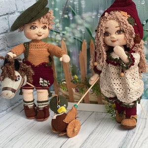 May include: Two crocheted dolls, one wearing a green hat and brown pants, the other wearing a red hooded dress, standing next to a wooden fence with a toy duck on wheels.