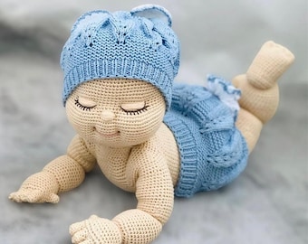 May include: A crocheted baby doll with a blue knitted hat and matching outfit. The doll has closed eyes and a sweet expression.