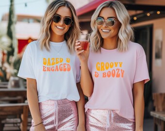 May include: Two women wearing sunglasses and smiling, each wearing a t-shirt with text. One shirt is white with "DAZED & ENGAGED" in orange and yellow. The other is pink with "GROOVY & BOOZY" in orange and yellow. Both are wearing sequin shorts.