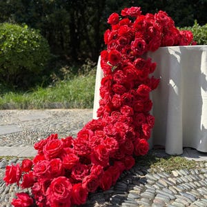 May include: A cascade of vibrant red roses spills from a white tablecloth, creating a dramatic floral display. The roses are densely packed, forming a lush, textured arrangement. The scene is set outdoors, with a stone path and greenery in the background.