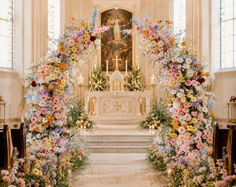 May include: A large, arched floral installation composed of a dense arrangement of pink, yellow, orange, white, and blue flowers with green foliage, framing a central aisle leading to a church altar with a religious painting and a chandelier.