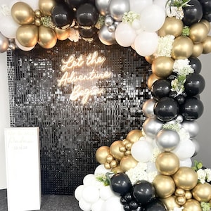 May include: A black, white, gold, and silver balloon arch with a neon sign that reads "Let the Adventure Begin". The balloons are arranged in a cascading pattern and are decorated with white flowers.