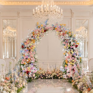 May include: A large, semi-circular floral archway constructed from a dense arrangement of multi-colored flowers including pink, yellow, blue, and white blossoms. The arch stands on a reflective floor, surrounded by matching floral floor arrangements.