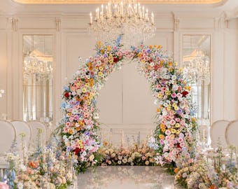 May include: A large, semi-circular floral archway constructed from a dense arrangement of multi-colored flowers including pink, yellow, blue, and white blossoms. The arch stands on a reflective floor, surrounded by matching floral floor arrangements.