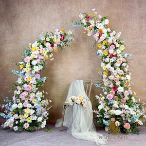 May include: Two floral arches, adorned with a variety of colorful flowers, create a romantic setting. A wooden chair draped with white fabric and a bouquet of flowers sits between the arches. The arches are perfect for weddings or special events.