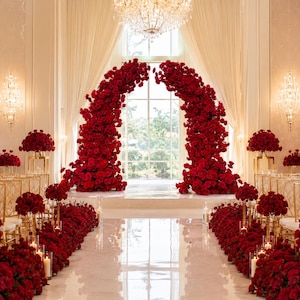 May include: A large, arched floral installation composed of deep red roses, positioned in front of a tall window. The aisle is lined with matching red rose arrangements on gold stands and floor-level clusters, accented by clear glass pillar candles.