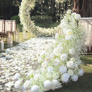 May include: A wedding ceremony setup with a floral arch and aisle. The arch is a large white floral ring. The aisle is covered in white flower petals. White roses and baby's breath are arranged in the foreground. The scene is outdoors on a sunny day.