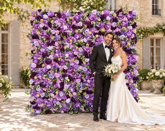 May include: A large, square floral backdrop wall featuring a dense arrangement of purple, violet, and lavender silk flowers, including roses and hydrangeas, standing outdoors on a stone patio in front of a light-colored stone building.