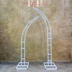 May include: White metal archway with two curved sections and a square base. The archway is made of metal tubing and is designed for use as a wedding or event backdrop.