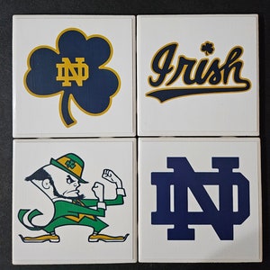 May include: Set of four white ceramic coasters with Notre Dame Fighting Irish logos. The coasters feature a green shamrock with the letters ND, the word "Irish" in a gold and blue script, a cartoon leprechaun, and the letters ND in blue.