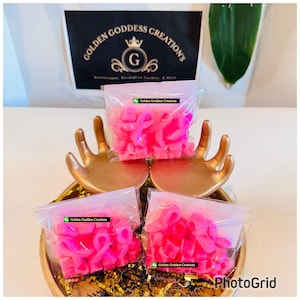 May include: A decorative display featuring small, pink ribbon-shaped wax melts in clear plastic bags. The bags are arranged in a gold-colored hand-shaped holder, with the text "Golden Goddess Creations" visible in the background.