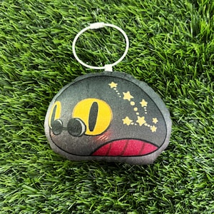 May include: A plush keychain featuring a cartoon cat face with large yellow eyes, black pupils, and a red mouth. The cat's head is dark gray with yellow star accents. A silver metal ring is attached.