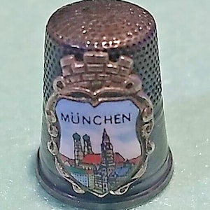 Peut inclure: Un dé à coudre gris foncé avec un écusson décoratif portant le mot "MÜNCHEN" et une illustration colorée d'un paysage urbain. Le dé à coudre a une surface texturée et un bord argenté.
