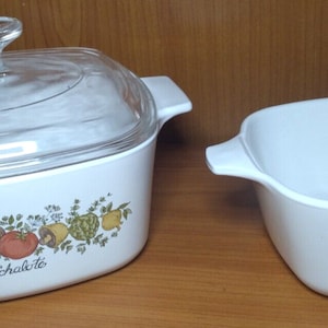 May include: Two white casserole dishes with a colorful vegetable design. One dish has a clear glass lid. The dishes have handles and are labeled "L'échalote" and "Le Persil".