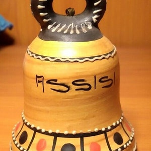 May include: A ceramic bell with a black and white handle and a brown, yellow and red painted design. The bell is decorated with white dots and has the word "Assisi" written on it.