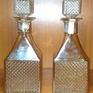 Op de afbeelding: Twee heldere glazen karaffen met vierkante, diamantpatroon stoppers. Het onderste deel van elke karaf heeft een textuur, diamantpatroon ontwerp. De karaffen hebben een rechthoekige vorm met schuine schouders.